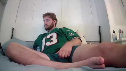 Media: Video of a bearded, light-skinned man in a green football jersey, sitting with legs spread on a bed, holding his crotch, in a dimly lit, sparsely furnished room.