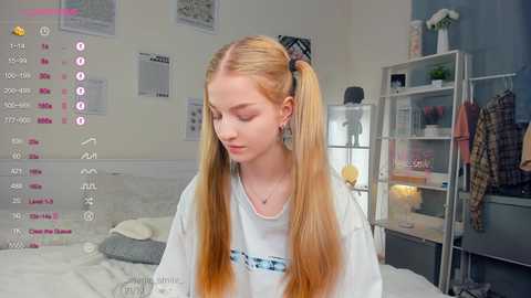 Media: A video of a young Caucasian girl with long, straight, blonde pigtails wearing a white T-shirt, standing in a modern, cluttered bedroom with a bed, shelves, and framed artwork.