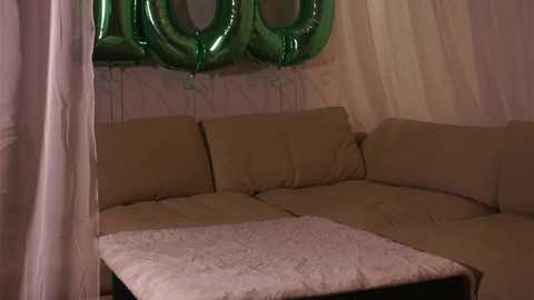 Media: Video of a beige sectional sofa with a plush white blanket, adorned with green balloons, against a pale pink curtain background.