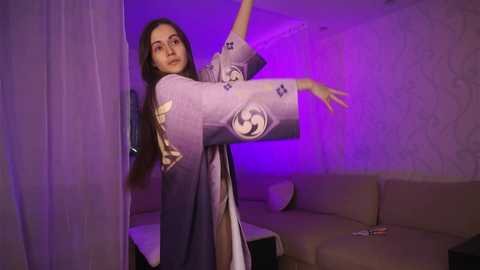 Media: Video of a woman in a long, flowing robe with floral patterns, dancing gracefully with outstretched arms under purple lighting. Background features a beige couch and textured white wall.