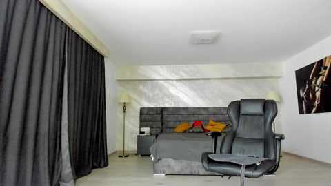 Media: Video of a minimalist, modern bedroom with a gray upholstered bed, black curtains, and a gray leather office chair; a black floor lamp stands beside the bed.