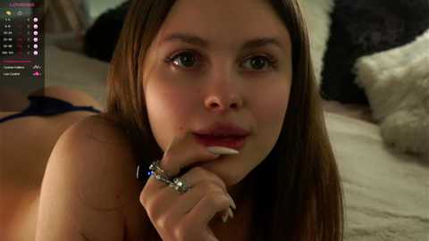 Media: A close-up video of a young woman with long brown hair, light skin, and full lips, lying on a bed with a fur blanket, wearing a blue bra, and holding a cigarette, with a video game interface in the top left corner.