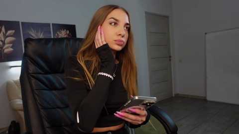 Media: Video of a young, light-skinned woman with long, straight brown hair, wearing a black crop top and headphones, sitting pensively on a black office chair in a modern, minimalist room with white walls and abstract art.