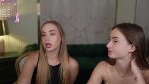 Media: Video of two young women sitting on a green couch in a dimly lit room. One has long blonde hair, wearing a black top, and the other has brown hair, wearing a gold necklace.