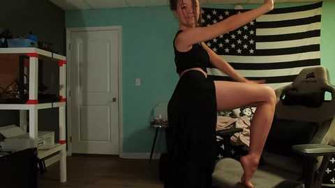 Media: Video of a fit woman with blonde hair in a black sports bra and pants performing a kick, in a bedroom with an American flag wall and gaming setup.