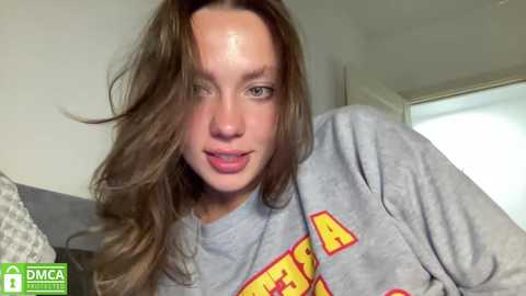 Media: Video of a young woman with long brown hair, wearing a grey shirt with red and yellow text, in a modern indoor setting with white walls and a window.