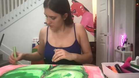 Media: Video of a woman with dark hair, wearing a blue tank top, painting a bright green and pink abstract flower on a canvas in a modern, brightly lit room.