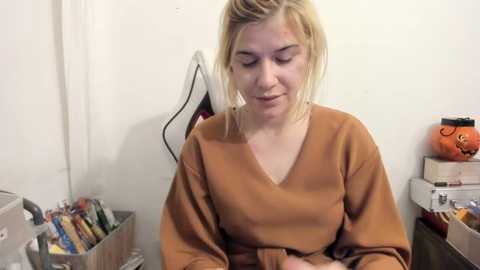 Media: Video of a blonde woman with fair skin in a mustard-colored robe, sitting in a cluttered room with shelves, boxes, and a stuffed toy.