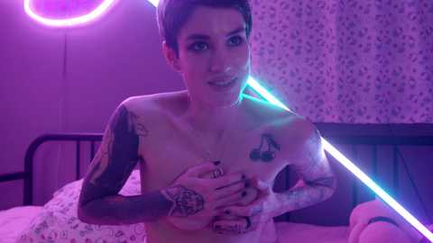 Media: Video of a topless, tattooed woman with short hair, holding her breasts, in a purple-lit bedroom with neon lights and floral wallpaper.