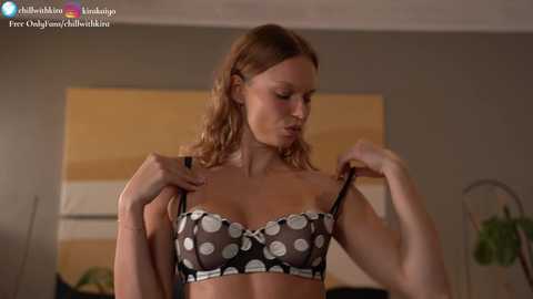 Media: Video of a slender, light-skinned woman with shoulder-length, wavy blonde hair, wearing a black and white polka dot bra, adjusting it in a dimly-lit room.