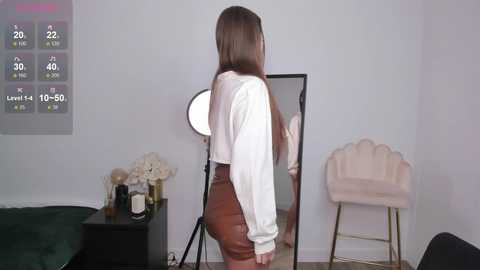 Media: Video of a young woman with straight brown hair, wearing a white long-sleeve shirt and brown leather shorts, standing in front of a large, light-colored wall-mounted mirror, with a digital clock showing 22:22.