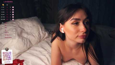 Media: Video of a young woman with straight black hair, wearing white earbuds, lying on a bed with white sheets, looking contemplative.