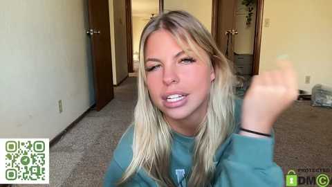 Media: A candid video of a blonde woman in a teal sweater, captured indoors on a carpeted floor. She appears to be speaking, with a distressed expression. The background features a beige wall, wooden doors, and a potted plant.