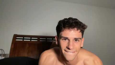 Media: Video of a shirtless, light-skinned man with short dark hair and a slight smile, standing in front of a wooden headboard.