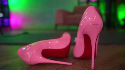 Media: Video of two glossy, bright pink high heels with stiletto heels, placed on a wooden floor. Background features a blurred green and purple light display, possibly a nightclub setting.