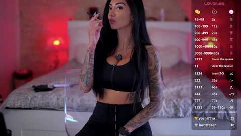 Media: Video of a tattooed, long-haired Latina woman in black crop top and pants, holding a cigarette, in a dimly lit bedroom with a bed and red lighting.