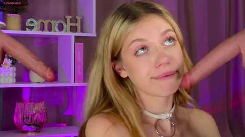 Media: Video of a blonde woman with blue eyes and fair skin, wearing a choker, looking up at two erect penises. Background includes shelves with \"SOMOH\" letters and books, under purple lighting.