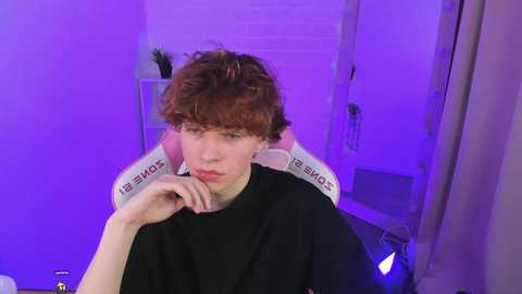 Media: Video of a young, pale-skinned man with curly red hair, wearing a black t-shirt, sitting in a white gaming chair under purple lighting, looking contemplative.