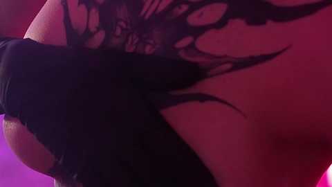 Media: Video of a close-up of a woman's exposed lower back, showing intricate black lace lingerie and tribal tattoo designs, against a pinkish-purple background.