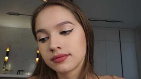 Media: Video of a young woman with fair skin and straight, light brown hair, wearing subtle makeup, including winged eyeliner, in a modern, well-lit room with white cabinets and a mirror with lights.