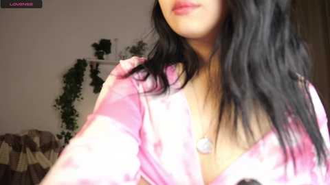 Media: Video of a woman with long, wavy black hair, fair skin, and wearing a pink satin robe. She is indoors, with a green plant and a bed visible in the background.