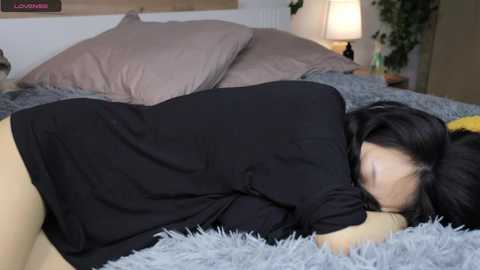 Media: Video of a woman with long black hair lying on a grey shaggy rug, wearing a black t-shirt, in a cozy bedroom with beige pillows and a lamp.