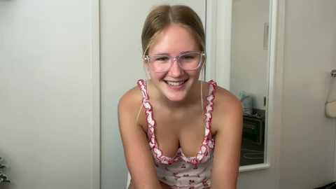 Media: Video of a smiling young woman with fair skin, light brown hair, wearing glasses, a floral-patterned tank top, and standing in a white-walled room with a mirror and door.