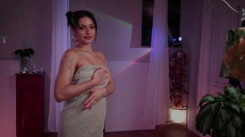 Media: Video of a slim, fair-skinned woman with dark hair in a bun, wrapped in a towel, standing in a dimly lit room with a floral-patterned lamp, a plant, and a vase on a table.