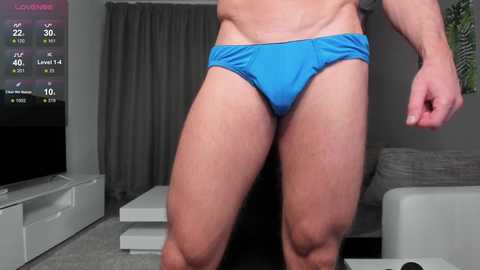 Media: Video of a muscular man in bright blue underwear, standing in a modern living room with grey curtains, white furniture, and a flat-screen TV displaying workout stats.