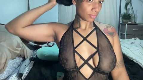 Media: Video of a young, light-skinned Black woman with short hair, wearing a black, crisscrossed halter top, revealing medium-sized breasts and tattoos on her right shoulder. Background includes a bed, pillows, and a dresser.