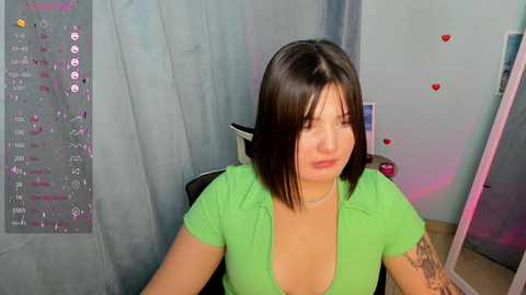 Media: Video of a young woman with shoulder-length dark hair, wearing a green shirt, sitting in a chair, against a soft, pastel-colored wall with heart decals.