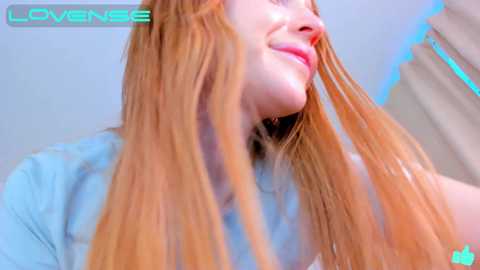 Media: A close-up video of a fair-skinned woman with long, straight red hair, smiling, wearing a light blue shirt, taken indoors with blue LED lighting.