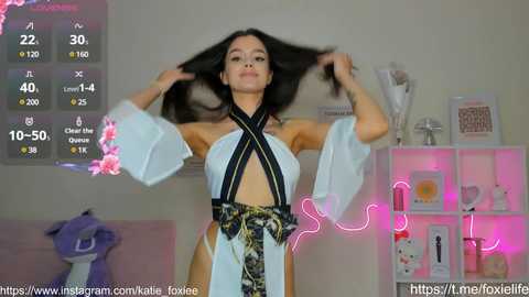 Media: A video of a young woman with long black hair, wearing a revealing, white and black patterned dress, posing confidently. The background features a white shelf with decorative items, pink neon lights, and a digital display showing her social media stats.