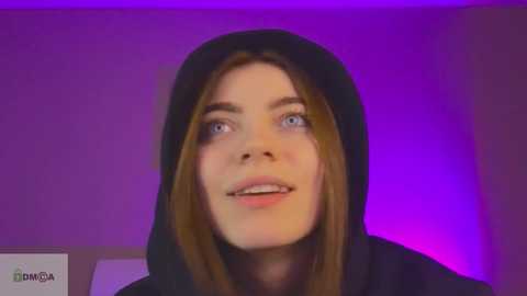 Media: Video of a young woman with fair skin and shoulder-length brown hair, wearing a black hoodie, smiling widely with blue eyes against a purple-lit background.