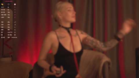 Media: Video of a white, slender woman with short, platinum blonde hair, wearing a black, plunging neckline top, sitting on a couch. Background features red curtains and dim lighting.