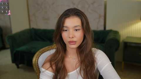 Media: Video of a young woman with long, wavy brown hair, fair skin, and full lips, wearing a white off-shoulder top, seated indoors. Background features a green velvet sofa, a lamp, and a patterned wall.