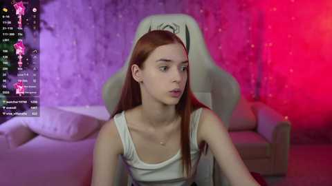 Media: A young, fair-skinned woman with long, straight red hair sits on a white chair in a dimly lit room. She wears a white tank top. The background features a purple and red light show.