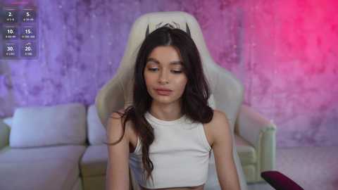 Media: A video of a young woman with long dark hair in a white crop top, sitting in a white gaming chair against a textured purple and pink background.