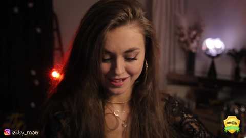 Media: Video of a young woman with long, straight brown hair, wearing a black lace top, smiling gently. Background features dim lighting and blurred decor.