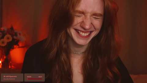 Media: Video of a smiling, freckled redhead woman with long, wavy hair, wearing a black top, against a warm, dimly-lit background with floral arrangements.