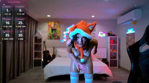 Media: A video of a woman in a fox costume with a Santa hat, standing provocatively in a modern, brightly lit bedroom with a bed, shelves, and neon lights.