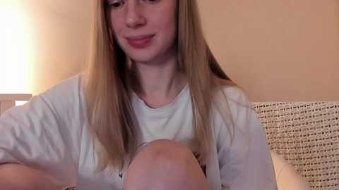 Media: A video shows a young Caucasian woman with long blonde hair and pale skin, wearing a white T-shirt, smiling while breastfeeding. The background features a beige wall and a white wicker chair.