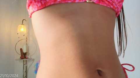 Media: Video of a slender, light-skinned woman wearing a pink tie-dye bikini top, with her midsection and toned abs visible. Background includes a lamp and white wall.