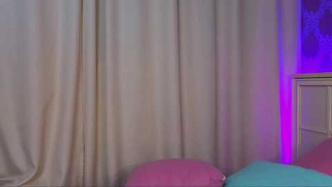 Media: A video of a child's bedroom with light beige curtains, a bed featuring pink and blue pillows, and a purple light illuminating the corner.