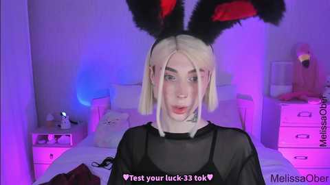 Media: Video of a young woman with platinum blonde hair, wearing black bunny ears, a sheer black top, and a white bed in a purple-lit room. Text at the bottom reads, \"Test your luck 35% tok <3.\