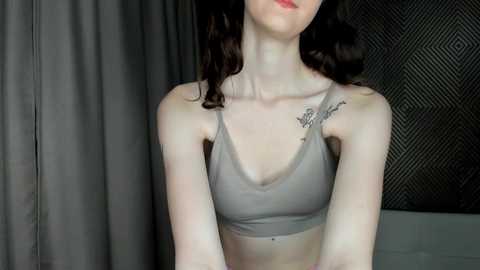 Media: Video of a slender, fair-skinned woman with dark wavy hair, wearing a gray sports bra, showing a small tattoo on her right shoulder. Background features dark curtains and a metal mesh wall.