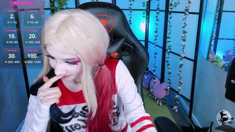 Media: Video of a young woman with long, platinum blonde hair, wearing a white and red Gotham City-themed shirt, sitting in a black gaming chair, biting her finger, in a dimly lit room with blue lighting and green vines.