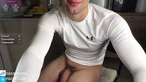 Media: A video of a young man with light skin and a slender build, wearing a white Under Armour long-sleeve shirt and no pants, sitting on a chair in a kitchen.