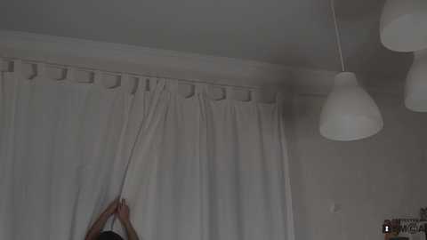 Media: A video of a person adjusting a white curtain with a hand, against a beige wall, featuring a hanging pendant lamp.