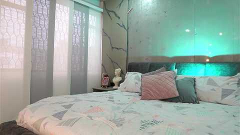 Media: A video of a modern, minimalist bedroom with a queen-sized bed covered in geometric-patterned bedding, adorned with pink and teal pillows. The wall features a large, mirrored headboard with a green backlit panel.